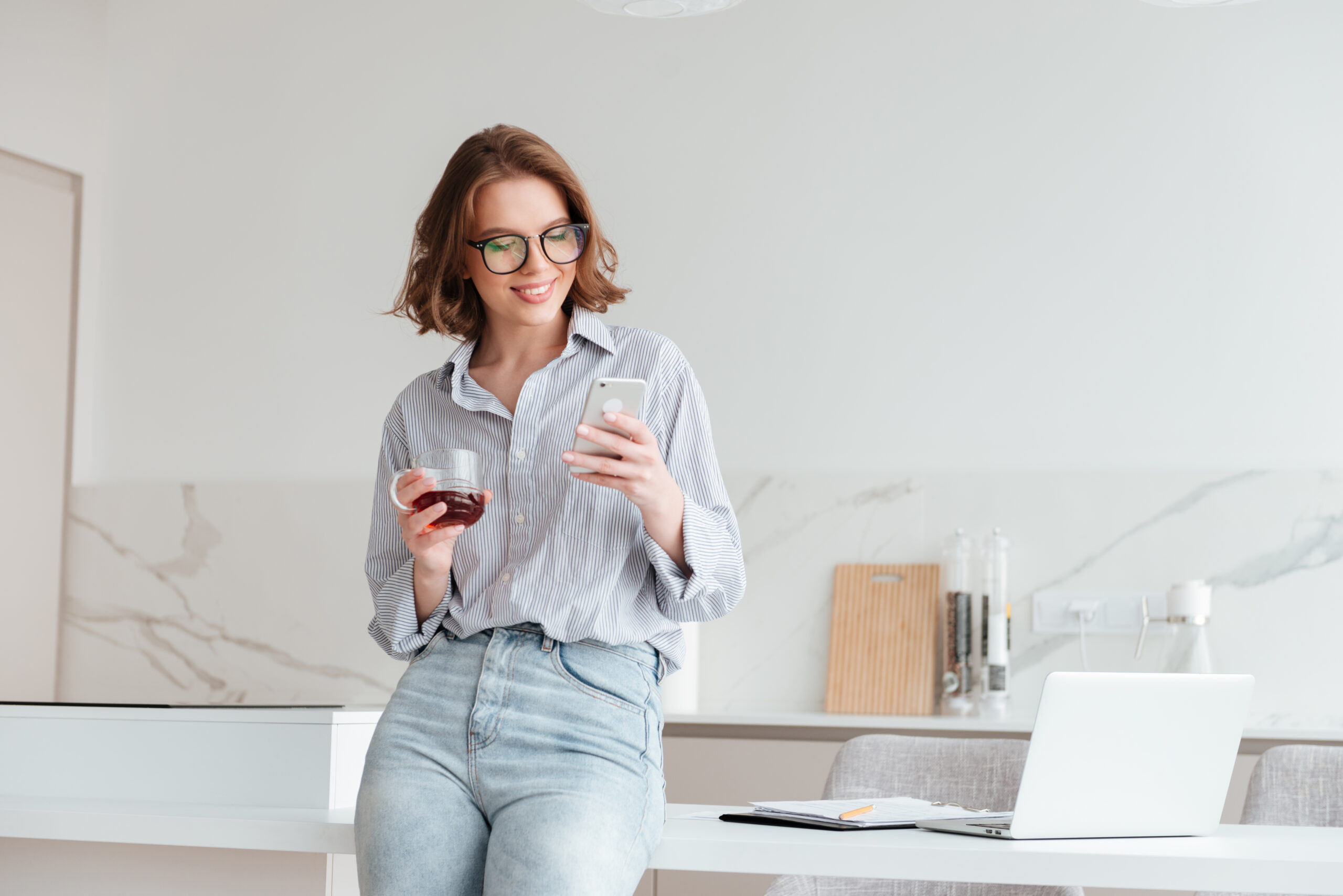 Portrait of a happy attractive woman using mobile phone while holding cup of tea and leaning on a table with laptop computer at home